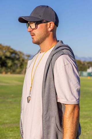Field coach observing practice wearing baseball cap, glasses and whistle on turf