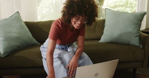 Smiling African American woman leaning toward laptop on green sofa, working from home