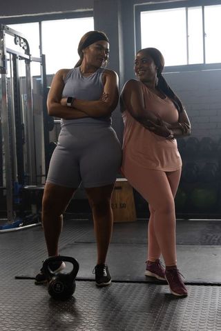 Two women standing with arms crossed in gym near kettlebell and weights showing fitness teamwork