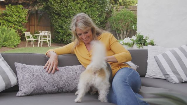 Woman enjoying playful time with her dog on an outdoor sofa in a beautiful garden. Ideal for themes related to pet care, outdoor living, relaxation, lifestyle, and human-animal bonding. Perfect for promoting home and garden furniture, dog-friendly spaces, and wellness products.