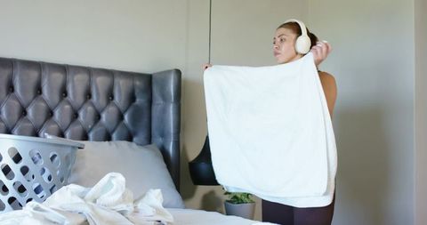 Woman folding white towel while wearing headphones in minimalist cozy bedroom