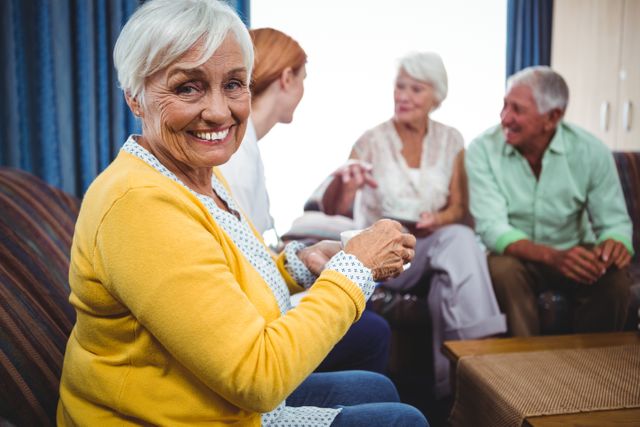 Senior woman smiling at camera while sitting with friends in a cozy living room. Ideal for depicting senior lifestyle, social gatherings, community living, and retirement activities. Perfect for use in advertisements, brochures, and websites related to elderly care, retirement homes, and social clubs.
