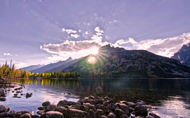 This picturesque scene captures a tranquil mountain lake at sunrise, with dramatic sunlight creating a captivating burst of light over a forested mountain range. Ideal for use in nature-focused content, travel magazines, environmental blogs, or inspiring background visuals.