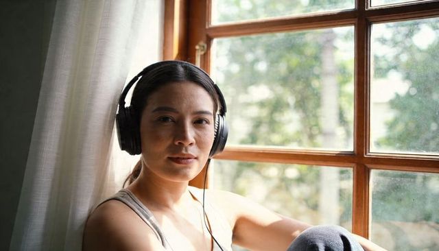 Woman listening with headphones on sunlit window seat in minimalist cozy home interior
