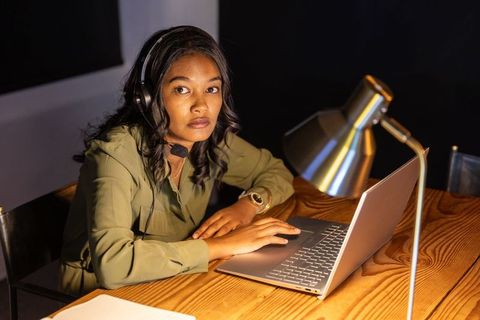African american woman wearing headset typing on laptop under desk lamp for remote work