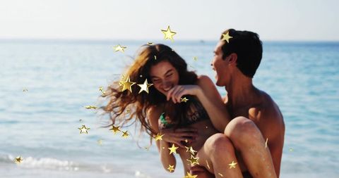 Couple laughing on sunlit beach while man carrying woman with golden star confetti