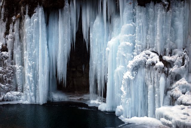 Beautiful frozen waterfall with large icicles creating a striking winter scene. The frozen water forms intricate ice formations, hanging dramatically over a partially frozen lake. Perfect visual for topics such as nature's beauty, winter, serene landscapes, and cold weather adventures.