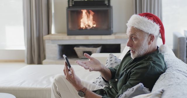 Elderly man wearing a Santa hat, sitting on a sofa, video calling friends or family. Fireplace glowing in the background creates warm, cozy atmosphere. Perfect for holiday greeting cards, virtual family gatherings, festive ads, and articles on modern technology connecting older adults.