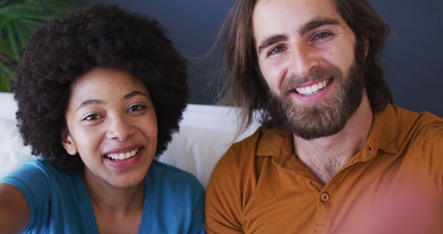 This image captures a happy interracial couple taking a selfie at home. It is suitable for promoting themes of love, diversity, happiness, and modern relationships. Ideal for use in advertisements, social media, blog posts, and articles about relationships, cultural diversity, and everyday life.