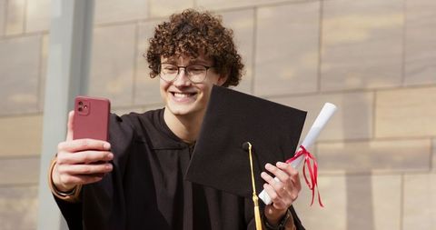 Smiling young graduate taking selfie with mortarboard and diploma outdoors