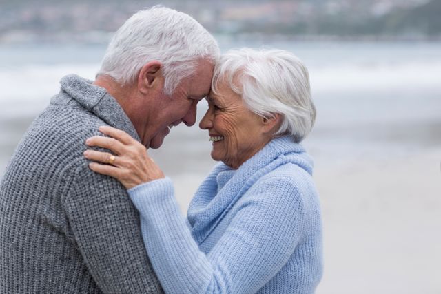 Elderly couple embracing and smiling on a beach, dressed in warm winter clothing. Ideal for use in advertisements or articles about senior romance, healthy aging, retirement lifestyle, and outdoor activities for seniors.