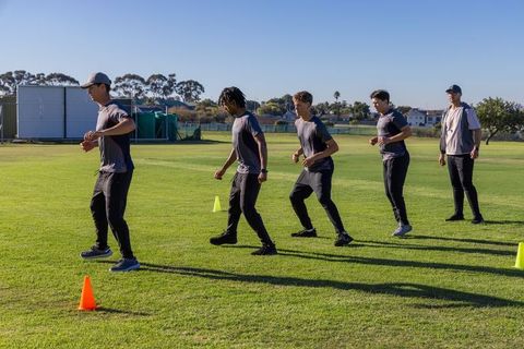 Diverse male athletes practicing cone footwork drill with coach timing on outdoor field
