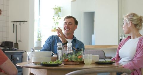 Friends laughing around dining table sharing salad in sunlit modern kitchen gathering