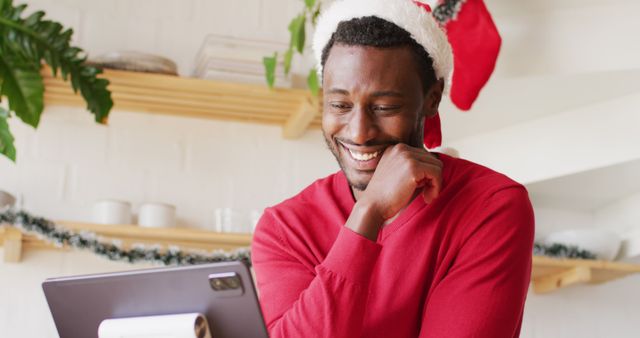 Man in Santa hat smiling while using tablet in festive setting. Ideal for holiday promotions, greeting cards, or blog posts about holiday cheer and technology use during festive season.