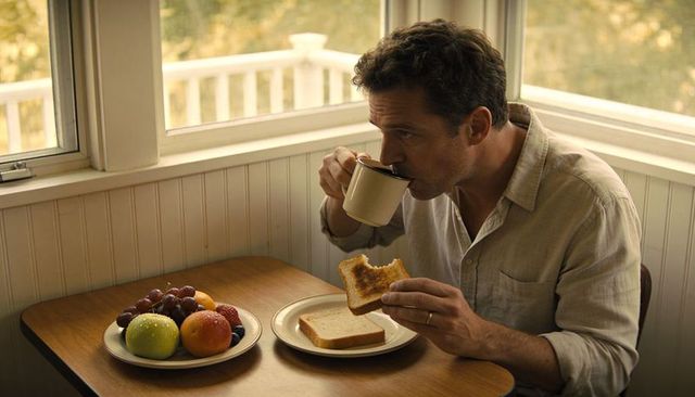 Man sipping coffee holding bitten toast in sunlit breakfast nook with fruit plate