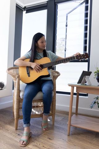 Asian woman tuning acoustic guitar in sunlit living room while following tablet lesson