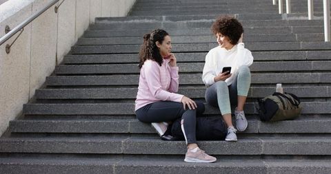 Two young women sitting on outdoor steps chatting and checking smartphone after workout