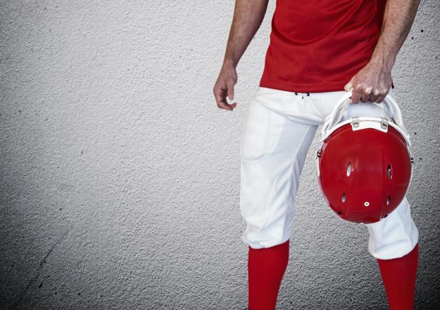 An athlete wearing a red and white football uniform is seen from the midsection down, holding a red helmet in one hand against a rough concrete wall. Ideal for use in sports-themed content, promoting athletic gear, or illustrating sportsmanship and teamwork in football.