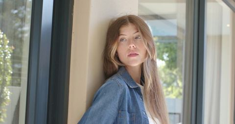 Young woman leaning by window in denim jacket looking contemplative in natural sunlight