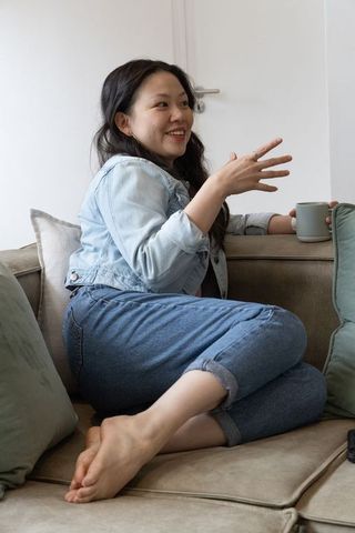 Asian woman relaxing on beige sofa holding mug and smiling while chatting in denim jacket