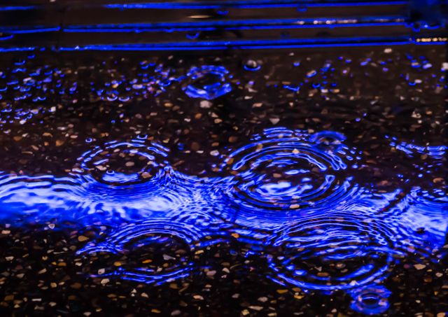 Glowing blue neon lights reflect on water surface with visible raindrops creating ripples. Ideal for concepts of night visuals, urban aesthetics, rainy weather, and abstract backgrounds. Useful in artistic projects, web design, and advertising materials.