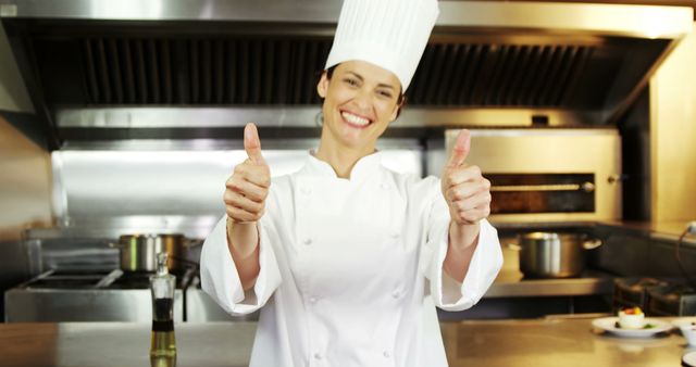 Female chef dressed in white uniform and hat showing approval with both thumbs up in a well-equipped commercial kitchen. Ideal for advertising culinary schools, cooking classes, restaurant promotions, and hospitality services. Demonstrates professionalism, positivity, and expertise in food preparation and culinary arts.