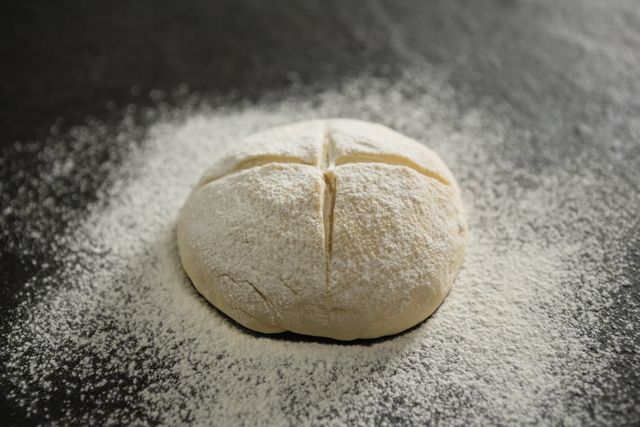 Unbaked bun dusted with flour on dark surface, perfect for illustrating baking processes, recipe blogs, or culinary articles. Ideal for use in food-related advertisements, cookbooks, or kitchen decor themes.