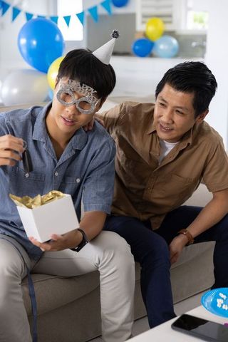 Father and teen son celebrating birthday at home opening gift with party masks