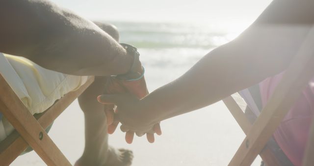 A couple is holding hands while sitting on folding chairs at a serene beach during sunset, capturing a tranquil and intimate vacation moment. Perfect for themes related to romance, relaxation, vacations, togetherness, and peaceful beach settings. Ideal for use in travel blogs, romantic getaway promotions, or lifestyle articles about spending quality time with loved ones at the beach.