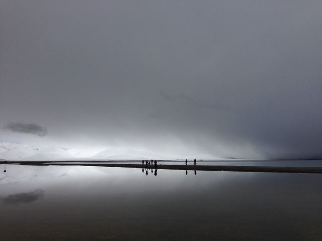 Image showcases a dramatic natural landscape featuring thick fog rolling over calm water. Silhouettes of a few people standing on a narrow strip of land bring a sense of scale and perspective. Reflection on the water creates a mirror effect, enhancing the serene atmosphere. Ideal for use in travel blogs, nature documentary visuals, meditation content, or art projects focusing on minimalism and vastness of nature.