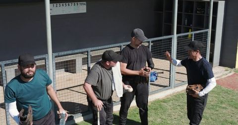 Baseball teammates chatting and preparing in dugout holding gloves, ball, bat and bottle