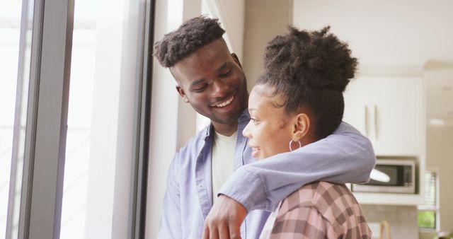 Image of happy african american couple standing at window and embracing. love, relationship, togetherness and spending quality time at home.