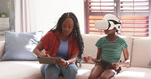 African mother using tablet while daughter wearing VR headset sitting on sunlit sofa