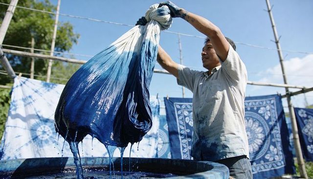 Artisan lifting indigo-dyed fabric from vat outdoors, dyeing blue cloth on bamboo rack