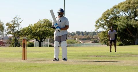 African american batsman batting on sunny park cricket pitch with fielder behind
