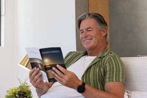 Mature man relaxing on sofa reading 'endless sunsets' magazine in green striped shirt