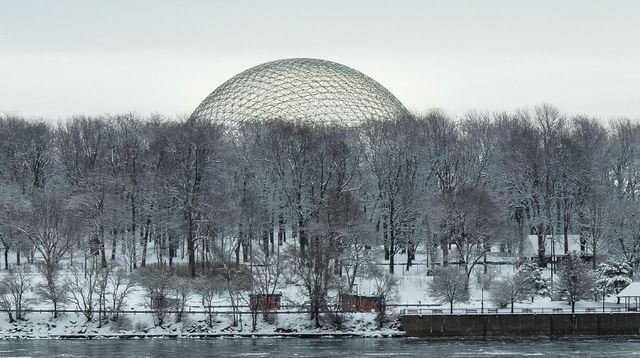 The image features a large geodesic dome set in a snowy landscape, surrounded by bare trees near a river. Salient against the sky, the dome's structured form adds contrast to the natural scene. This could be used for showcasing architectural designs, travel promotion during winter, or nature-themed projects.