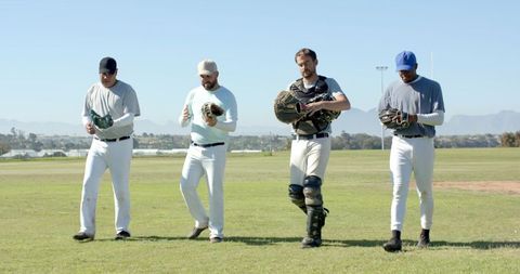 Four Baseball Players Walking Across Sunlit Outfield Carrying Gloves and Catcher Gear