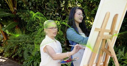 Senior art mentor guiding young student painting outdoors at easel in lush garden