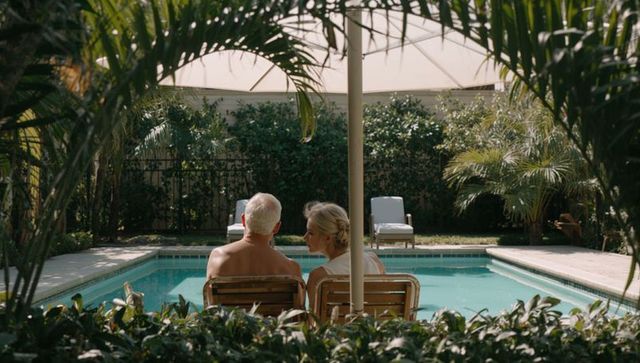 Senior couple relaxing by private tropical backyard pool under umbrella, wooden chairs