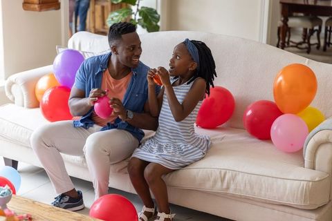 Smiling father and daughter inflating colorful balloons on sofa for home birthday party
