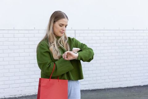 Woman checking smartwatch while holding coffee and red shopping bag by white brick wall