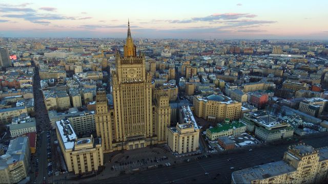 Aerial drone view of Moscow at sunset. Central focus on an iconic Stalinist skyscraper with golden light reflecting off the buildings. Snow patches visible, contributing to the winter ambiance. Ideal for use in travel articles, architectural studies, and city tourism promotions.
