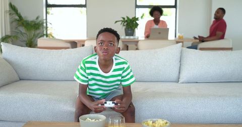 African american boy playing video game while parents working in open living room