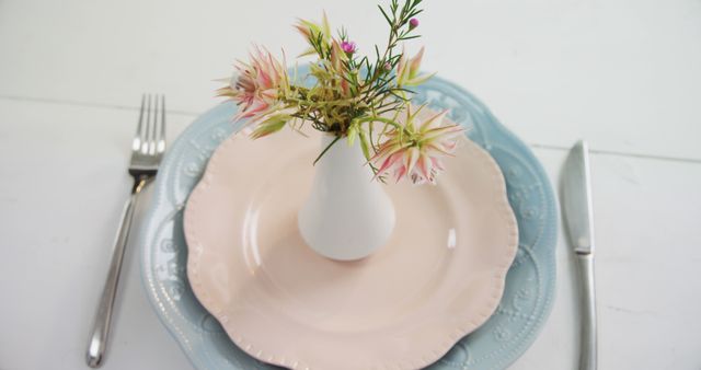 A simple and elegant table setting features a pink plate with a small vase of flowers, flanked by silverware, with copy space. The arrangement suggests a fresh, springtime vibe ideal for a brunch or casual dining experience.