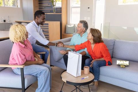 Diverse couples greeting and shaking hands in sunlit modern living room with gift bag