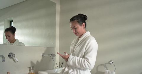 Mid-adult asian woman applying moisturizer at minimalist bathroom vanity wearing white robe