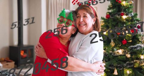 Hugging seniors wearing christmas tree glasses by decorated tree and cozy fireplace