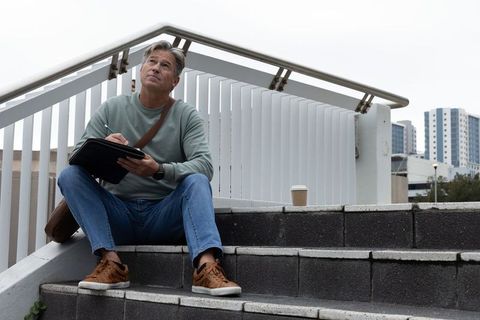 Middle-aged man sitting on urban steps writing in leather folio with takeaway coffee