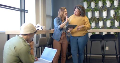 Diverse coworkers collaborating in modern office lounge pointing at tablet and laptop